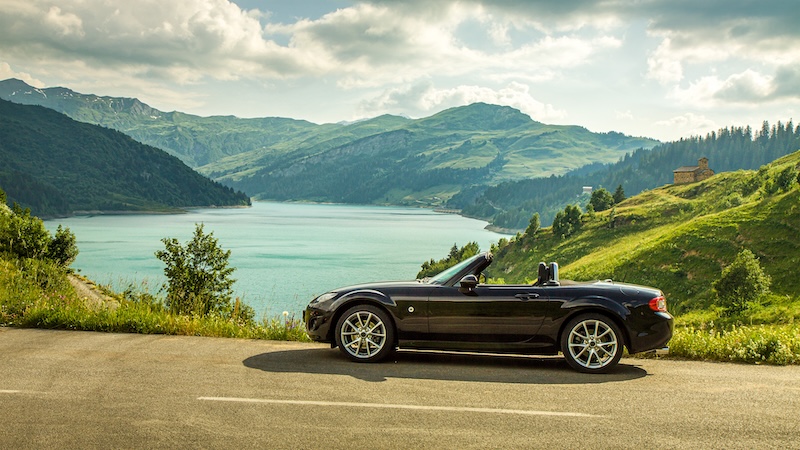 A black Mazda Miata convertible parked by a scenic mountain lake under a sunny sky, showcasing its sleek design. Image used for editorial purposes.